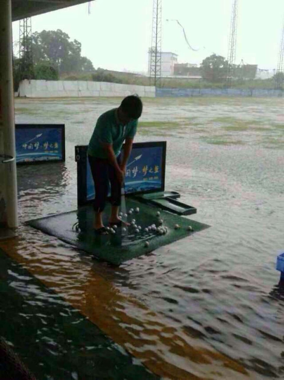 果岭被淹，沙坑失形，一位身着蓝色上衣的球友正凝神准备挥杆，脚下是漫过草皮的积水，倒映着阴沉的天空。