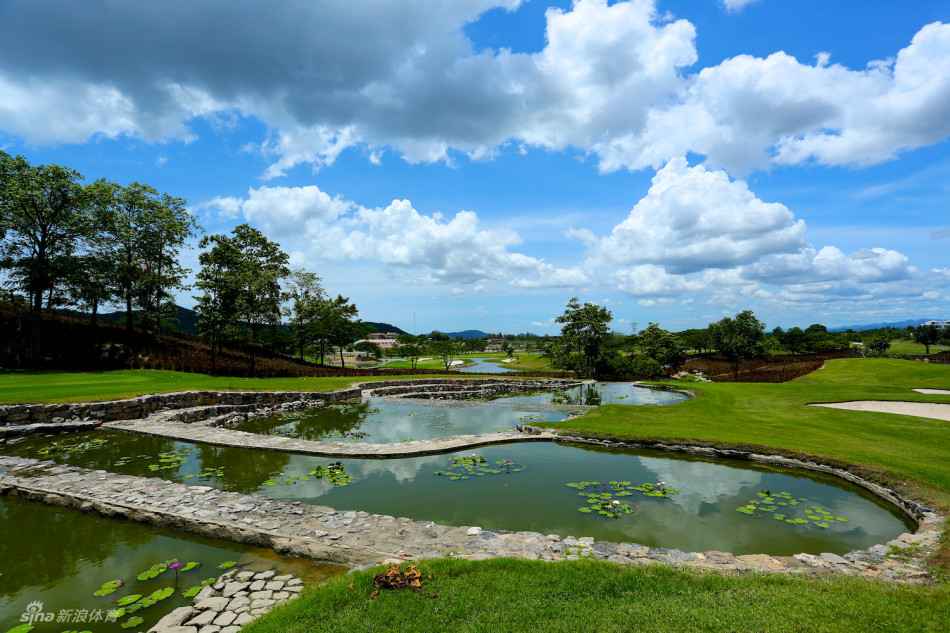 阳光洒落在宽阔的水障碍上，波光粼粼的水面倒映着蓝天白云，为秀美的球场景观增添了静谧而危险的元素