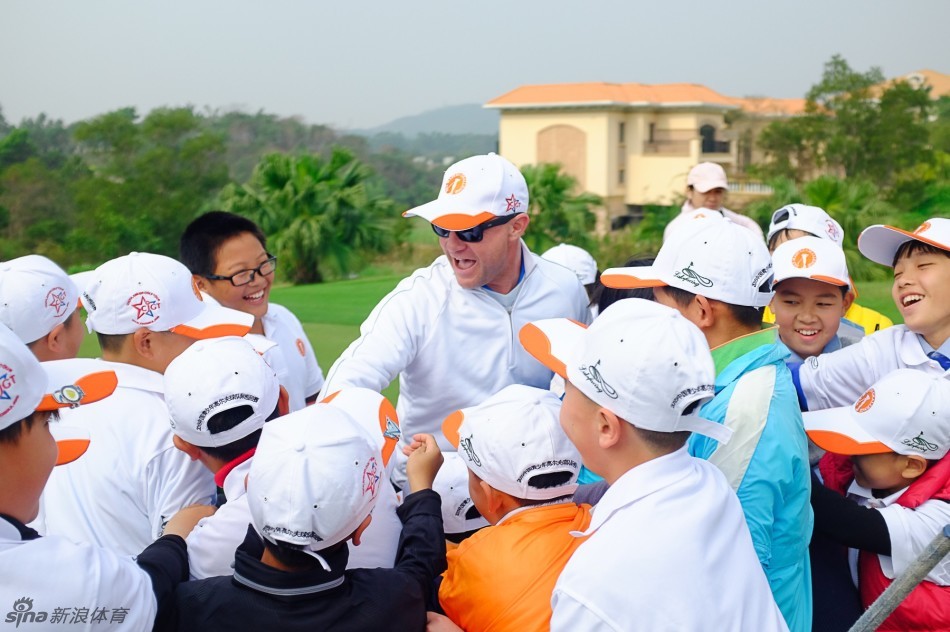 金发美国教练在球道中央，正微笑着指导一位中国少年调整挥杆姿势，阳光将他们的身影拉长