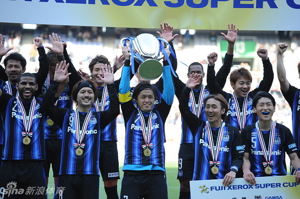 大阪钢巴队长在漫天金带中高举2026超级杯冠军奖杯，目光坚毅，身旁队友疯狂簇拥，喜悦定格在这一刻。