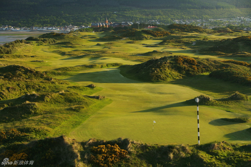 从高处俯瞰皇家邓恩郡第4洞果岭，蜿蜒的沙坑如自然雕刻的艺术品守护着目标，远方默恩山脉的轮廓在光影中若隐若现