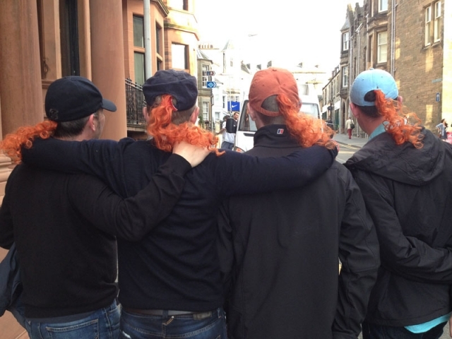 几位球迷身穿防水外套，聚集在临时搭起的露天棚下，手里举着满满的啤酒杯正在碰杯，表情轻松愉悦，背景是典型的苏格兰式建筑。