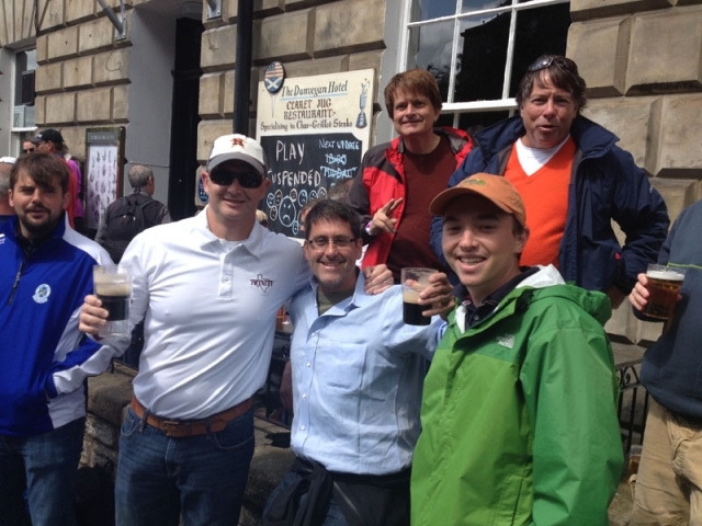 两名球迷并肩站在户外，其中一人正专注地用手机拍摄球场或同伴，另一人则面带微笑望向镜头，手中透明的塑料啤酒杯里酒液摇曳。