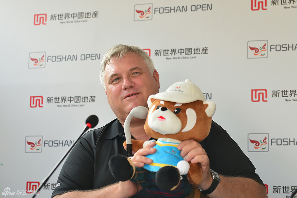 佛山高尔夫球会总经理韩祈福双手托举着吉祥物豆豆，亲切地向观众展示，背景是赛事主题板，现场气氛融洽。