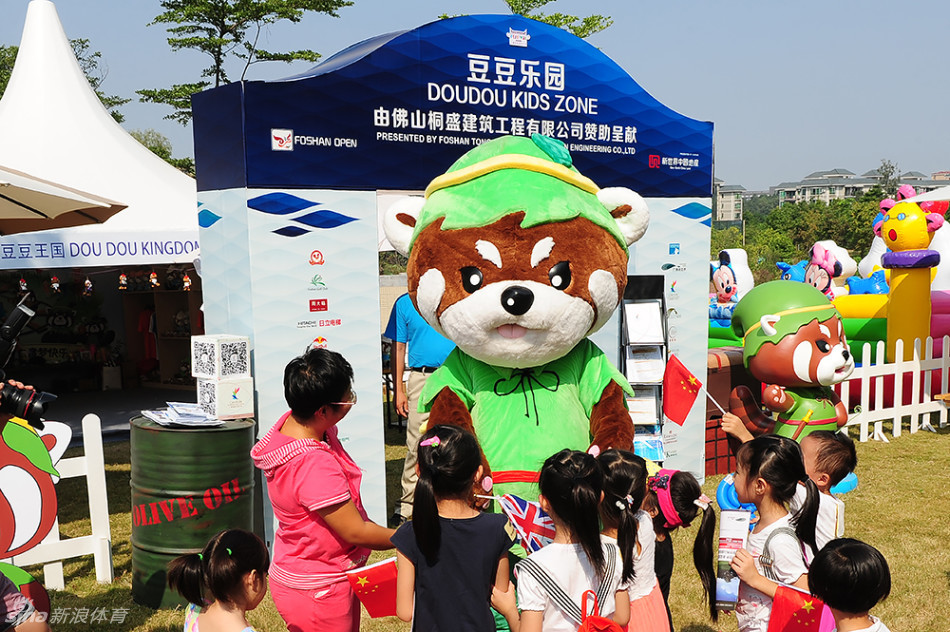 吉祥物豆豆蹲下身来,正与一个满脸笑容的小女孩击掌互动,阳光洒在他们身上,画面温馨动人