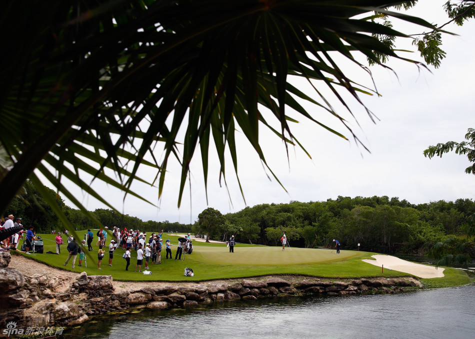 广阔湖畔果岭全景，雨丝如织，几名球员色彩鲜明的服装在灰绿背景下宛若移动的焦点