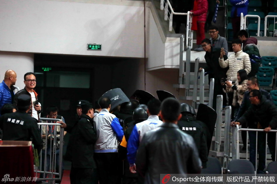在安保人员的层层护卫下，裁判员低着头快步疾走，试图快速穿过球员通道入口，身后的背景是模糊不清但又充满压迫感的看台人群。