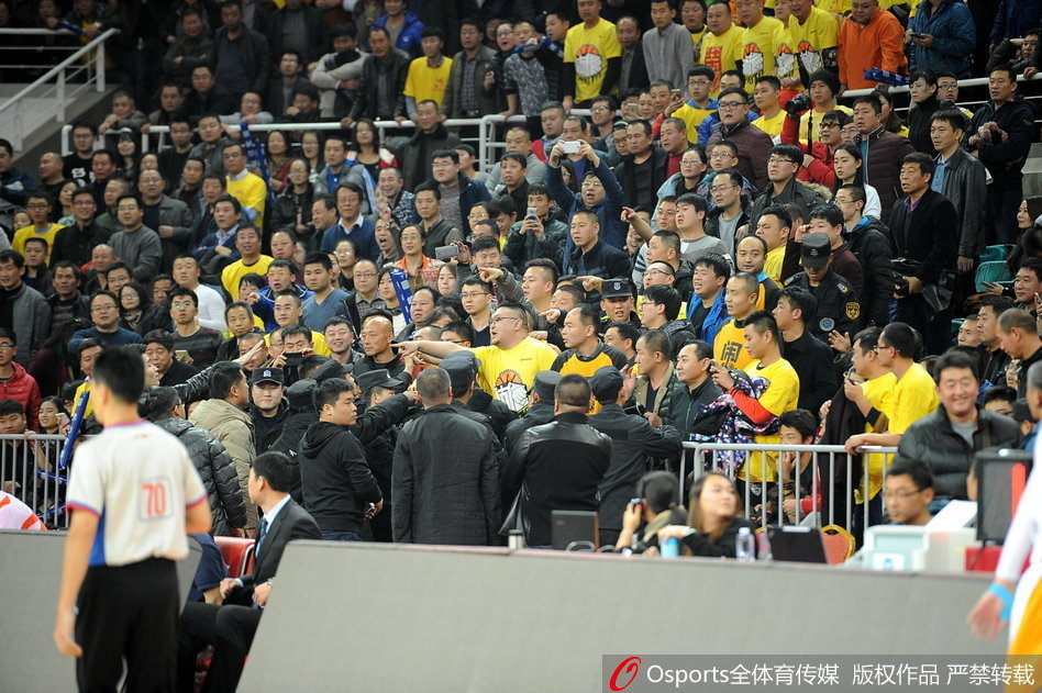 看台上的球迷们集体站立，目光齐刷刷地投向场地一角的冲突区域，许多人高举着双臂，面部表情因高声呼喊而变得扭曲，现场气氛紧张到了极点。