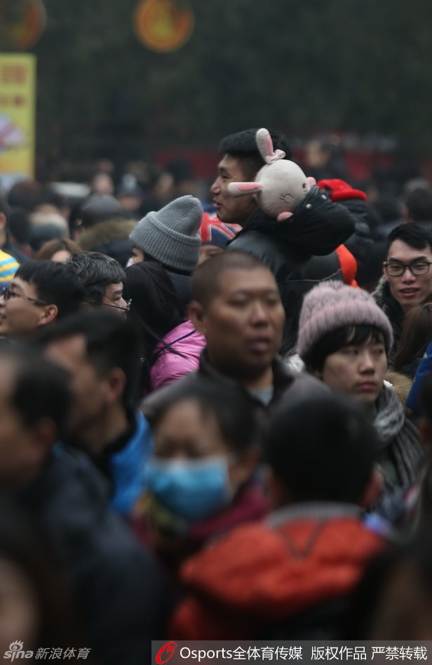 常林正全神贯注地参与庙会上的趣味游戏，他怀中抱着一个巨大的卡通毛绒玩偶，显然是刚刚获得的战利品，脸上带着轻松愉悦的笑容，尽显硬汉柔情。