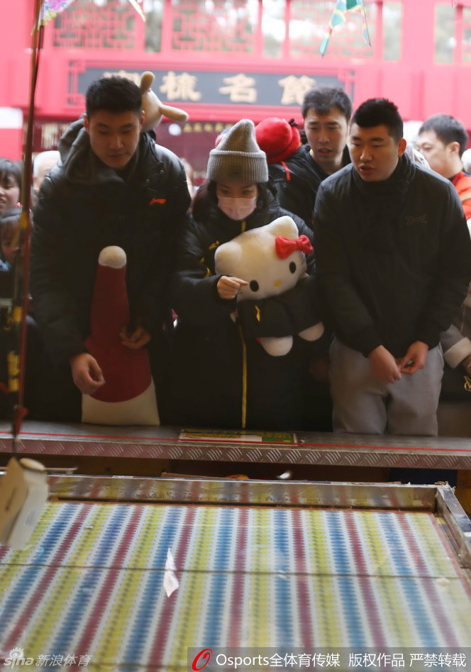 逛累了的常林与女伴在庙会休息区小憩，他手中依然抱着那个大玩偶，脸上带着些许疲惫却十分满足的神情，身后是庙会熙熙攘攘的人流和喜庆的装饰。