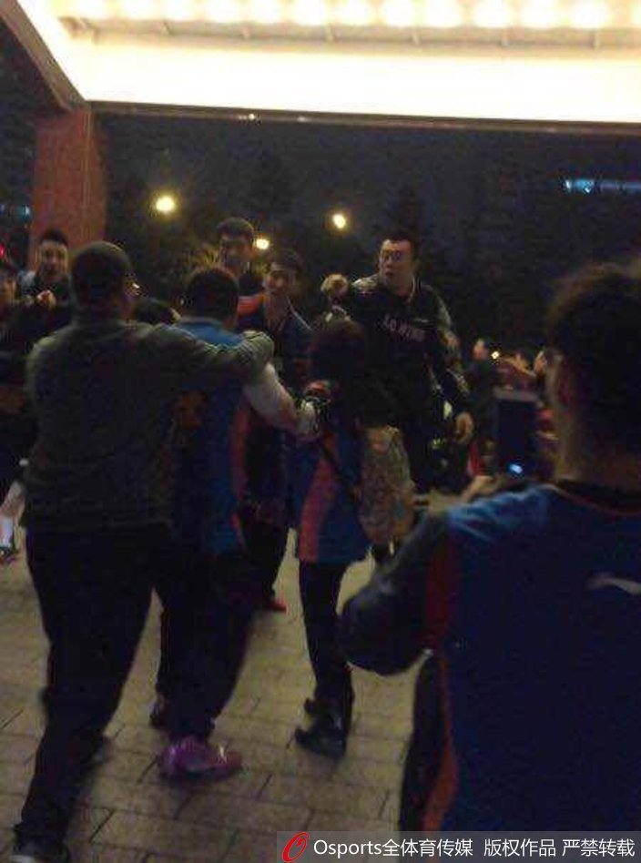 深夜的酒店门口一片混乱，多名身穿制服的人员试图挤进人群中心拉开斗殴双方，外围的球迷仍在举着手机拍摄，闪光灯与喧哗声交织。