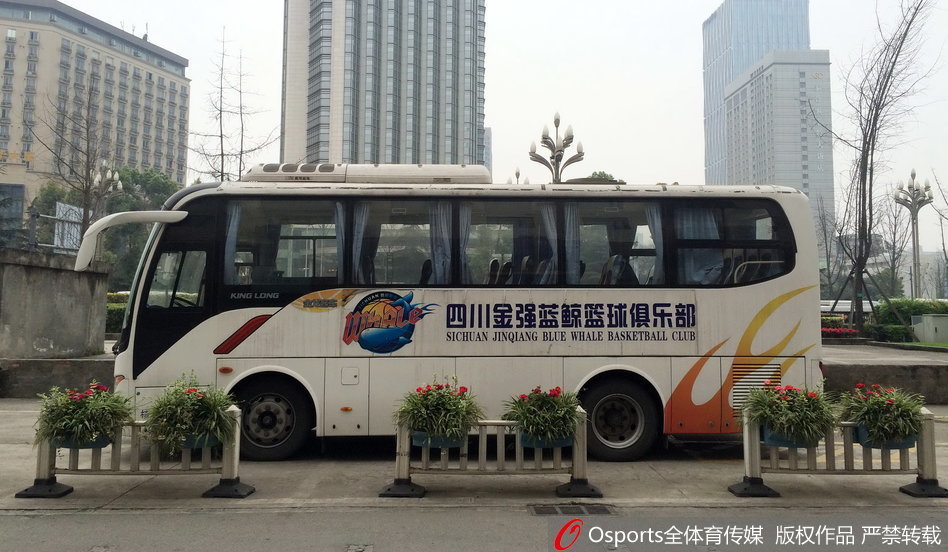 空旷的酒店门前，一辆喷涂着‘四川队’标识的蓝白大巴车孤零零地停靠在路边，车身反射着午后惨白的阳光，车内空无一人，仿佛在诉说着赛事停摆的紧张与不确定。