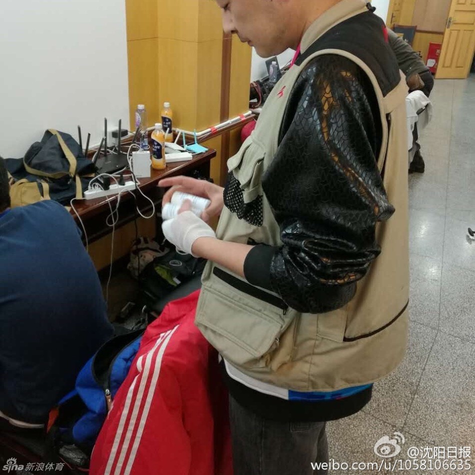 媒体工作室里，一位年纪较长的记者正低头看着手机，腕间的绷带在衣袖旁若隐若现。周围年轻的同行们同样手缠绷带，各自忙碌，整个空间弥漫着一种静谧而坚定的职业氛围。