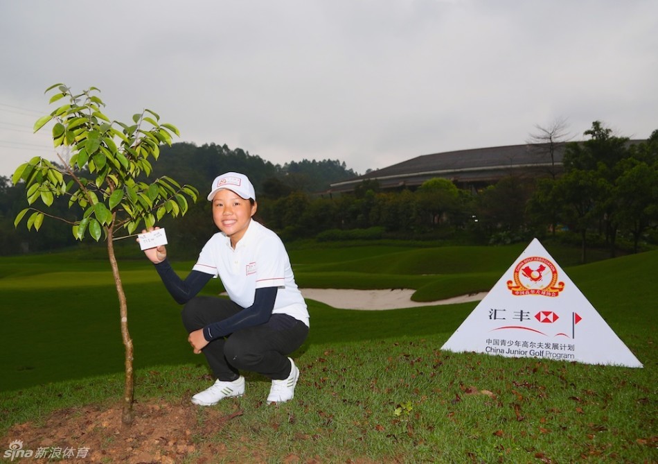 一位小球员正将一张写有祝愿的爱心形状心愿牌，仔细地系在新栽种小树的枝桠上。