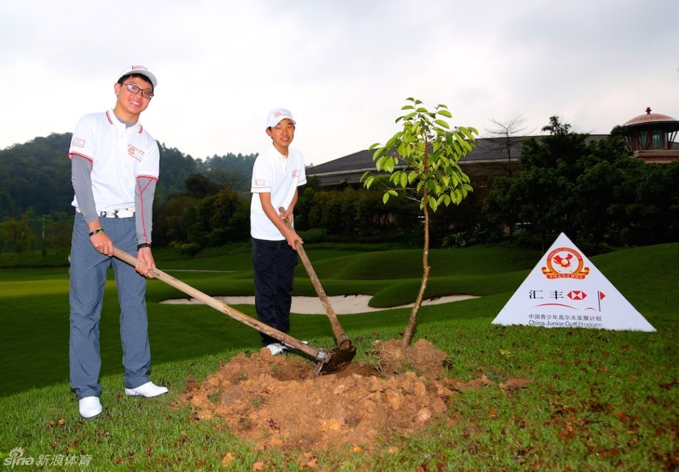 小球员梁恩旗手持铁锨，面带微笑，正用力将泥土铲入树坑，专注而投入。