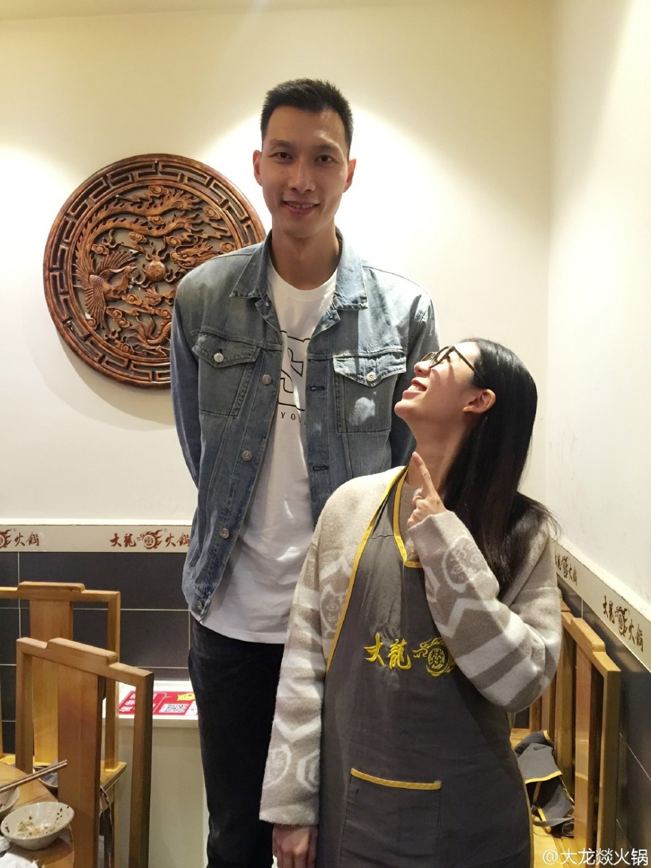 易建联穿着休闲深色T恤，在火锅店内微微弯腰侧身，满脸笑容地与身边身穿红色工作服的女店员合影。女店员兴奋地举起手掌，阿联则配合地伸出宽大的手掌与其紧紧相贴，巨大的身高差让这一幕显得格外有爱。