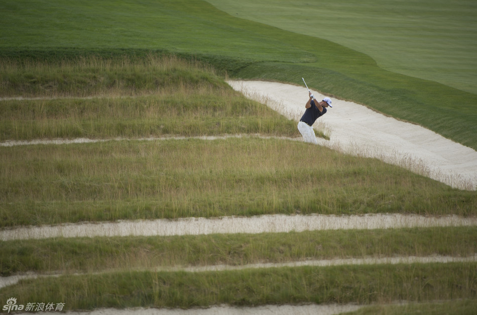 瑞奇·福勒在沙坑中奋力一击，沙粒飞扬，其鲜艳的着装与奥克芒深绿的背景形成强烈对比，诠释着激情与传统的碰撞。