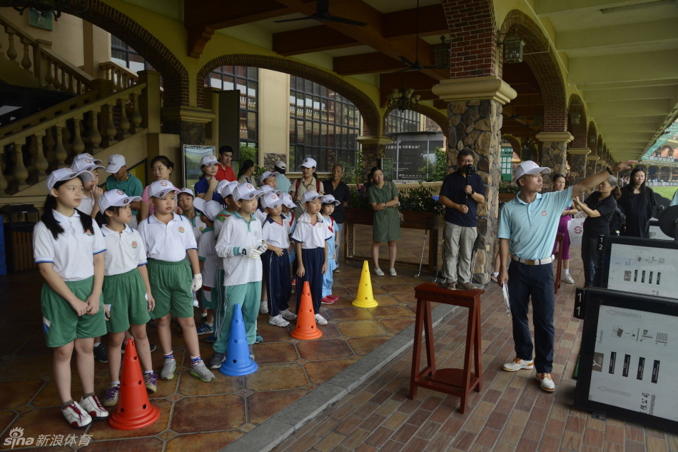 教练站在孩子们中间讲解比赛规则，手指向远方果岭，学生们仰头倾听，构成一幅生动的教学图景