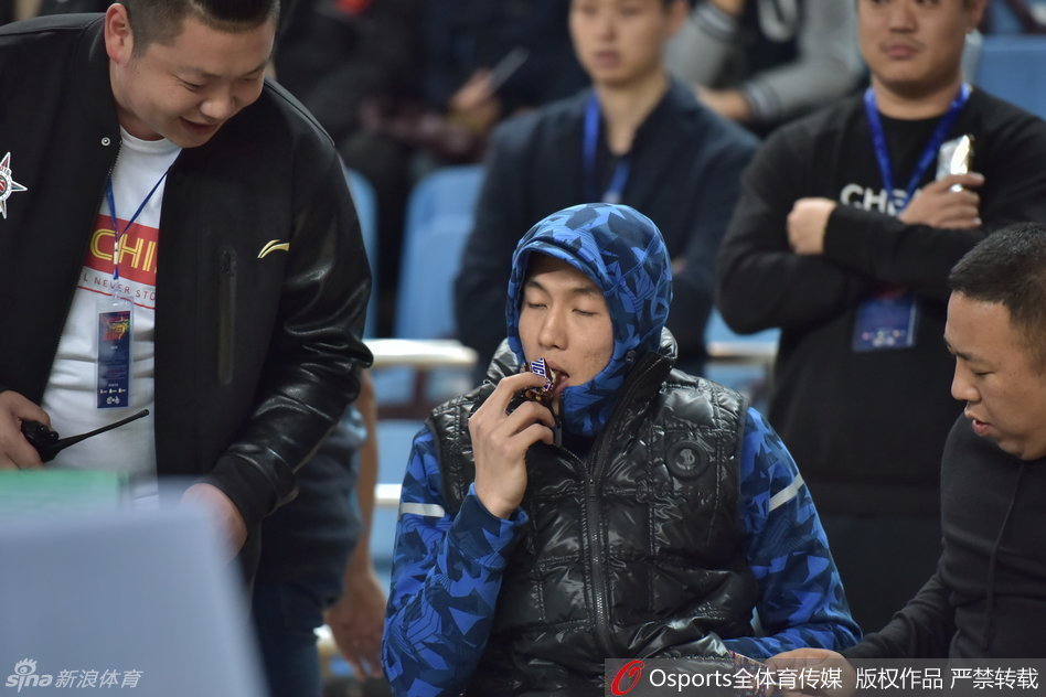 郭艾伦站起身在场边活动，厚实的外套显得他有些笨拙可爱，他依然不忘拿着那根棒棒糖，仿佛这是他在这个寒冷秋日里最重要的伙伴，脸上写满了“心情不错”。