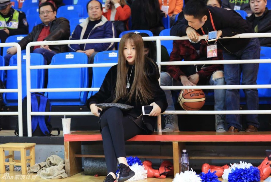 一位戴着眼镜的八一队铁杆女球迷静静坐在场边观战，她面容清秀，眼神专注地望向赛场，温柔的气质与场上激烈的对抗形成鲜明对比。
