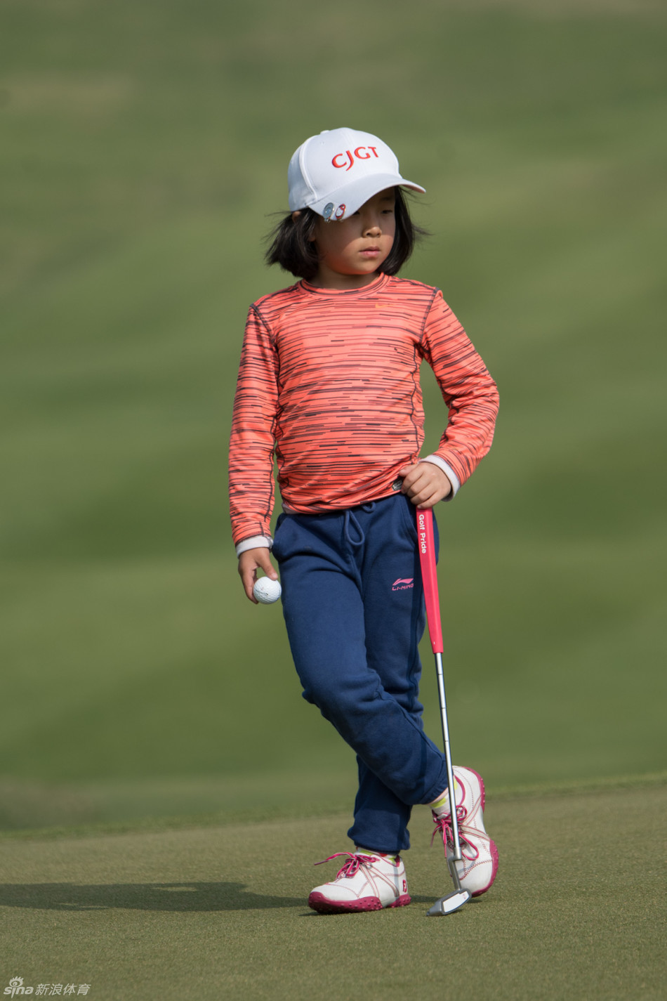 小女孩在果岭旁专注地审视地形，阳光洒在她认真的脸庞上，仿佛正在心中规划着小球进洞的完美弧线。