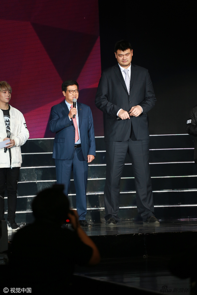 姚明与现场的音乐人及嘉宾并肩而立，巨大的身高差构成一幅有趣的画面，他正侧耳倾听，眼神中流露出对音乐世界的尊重与好奇。