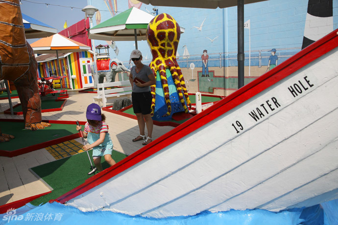 海滨小镇旁一个宁静的迷你高尔夫球道,背景是蓝天与远处的摩天轮,色彩明艳的障碍物在阳光下显得格外可爱。