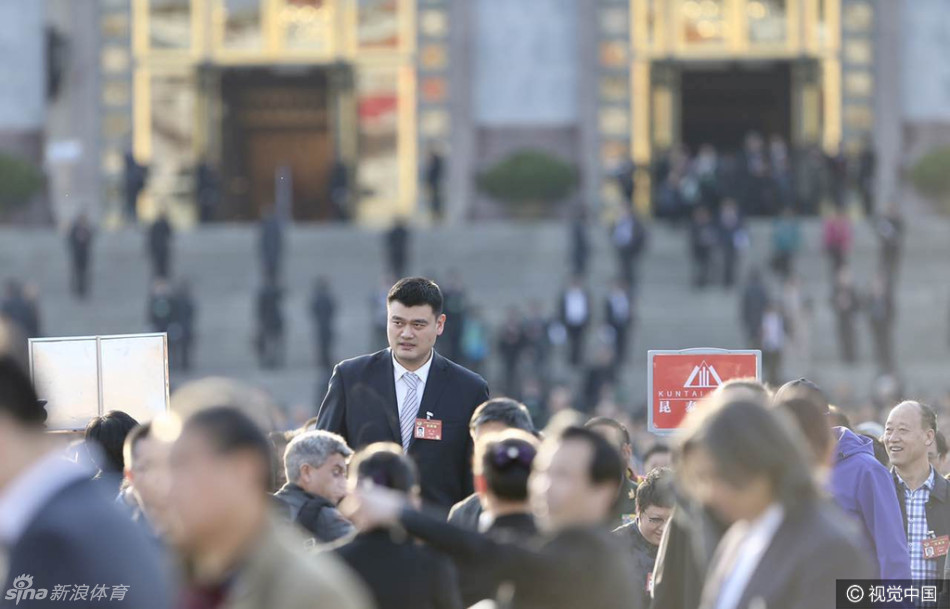 姚明独自行走在大会堂外的台阶上，背景是庄严的国徽与飘扬的红旗，他以仰望的姿态看向远方，仿佛在审视着体育强国的未来蓝图