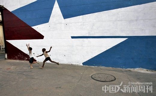古巴哈瓦那街头一面画有国旗的墙体旁，两名儿童正在嬉戏。在古巴的许多街头，常可以见到各式各样的墙画，内容多为国旗、革命主要领导人形象等。