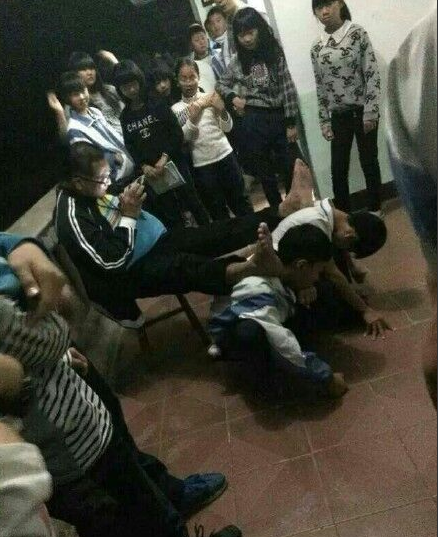 一名较胖的男子坐在椅子上玩着手机，赤脚分别放在两孩子的后背上。